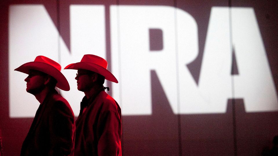 Zwei Männer mit Cowboyhüten stehen vor dem Logo der NRA