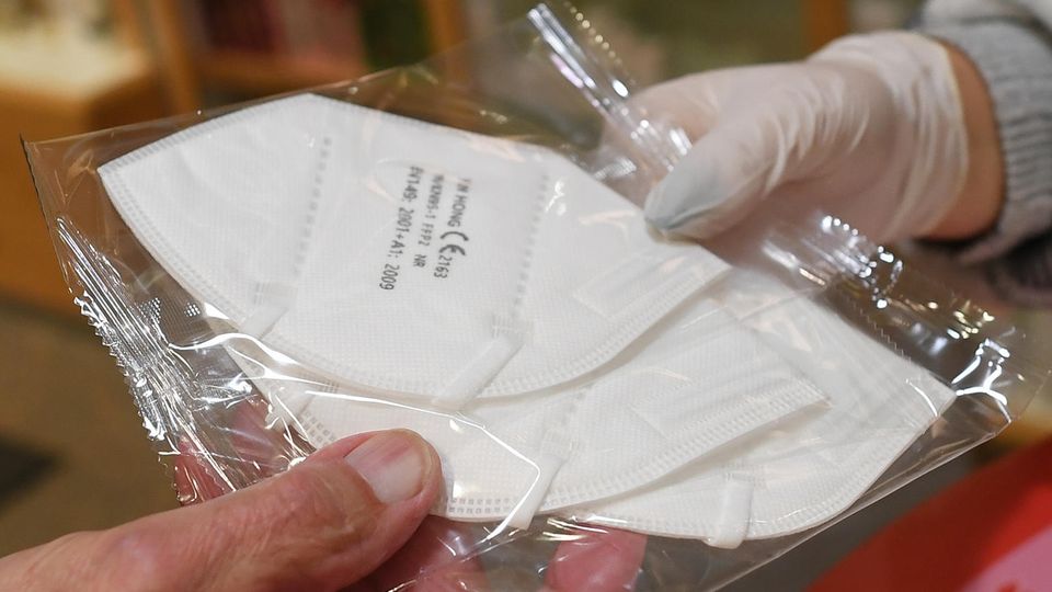 Eine Hand im Gummihandschuh hält eine Klarsichtfolie mit drei FFP2-Masken, die alte Hand eines Mannes nimmt sie entgegen