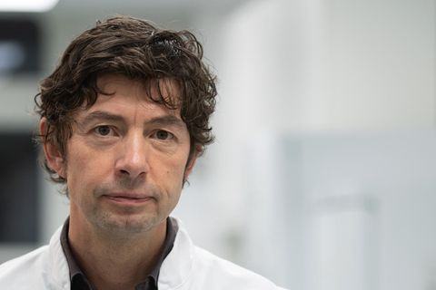 Ein Mann mit braunen Locken und im weißen Arzt-Kittel steht auf einem Klinik-Flur