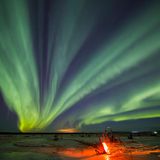 Grün und orange: Wie breite Kondensstreifen wirken hier die Nordlichter am Nachthimmel, während ein Lagerfeuer für einen farblichen Kontrast und Wärme sorgt.