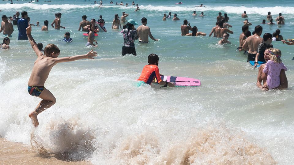 Australien Sonne Strand viele Menschen Wasser Welle Eine etwa achtjähriger Junge in Badeshorts springt am Strand über eine brechende Welle. Im Wasser sind schon viele Erwachsene