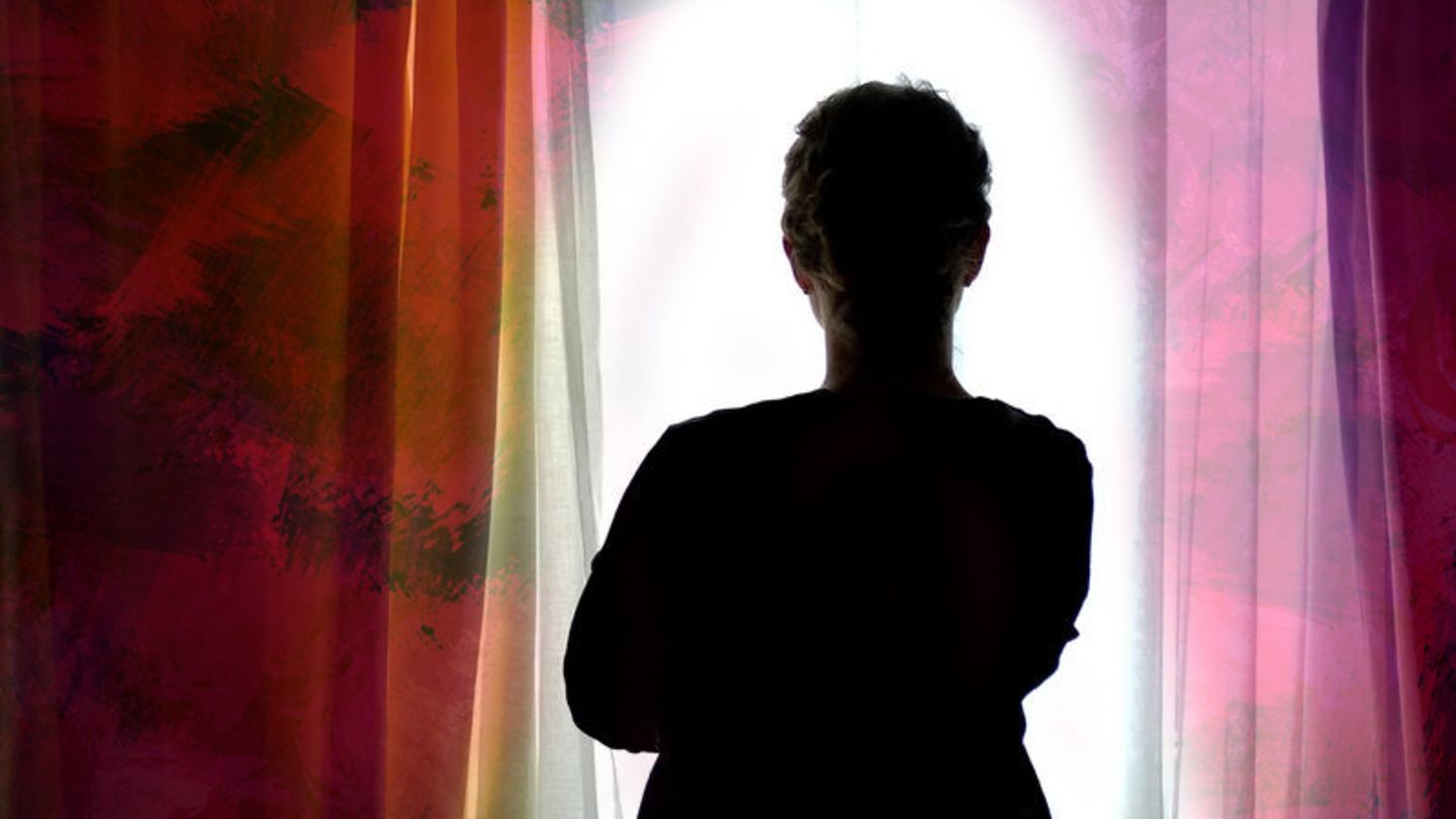 Eine Frau steht vor dem Fenster und schaut hinaus, man sieht sie von hinten im Schatten