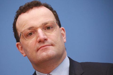 Vor einer blauen Wand legt Bundesgesundheitsminister Jens Spahn seinen Kopf schräg. Er hat braune Locken, trägt Anzug und Brille