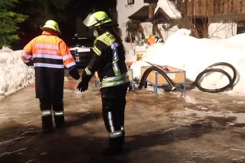Zwei Feuerwehrleute stehen auf einer Straße bis zu den Knöcheln im Hochwasser. Hinter ihnen ein hoher Schneehaufen