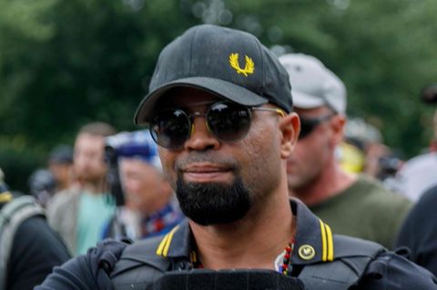 Ein Mann mit schwarzen Basecap, Sonnenbrille und schwarzem Polo-Hemd mit gelbem Streifen am Kragen steht vor einer Demo