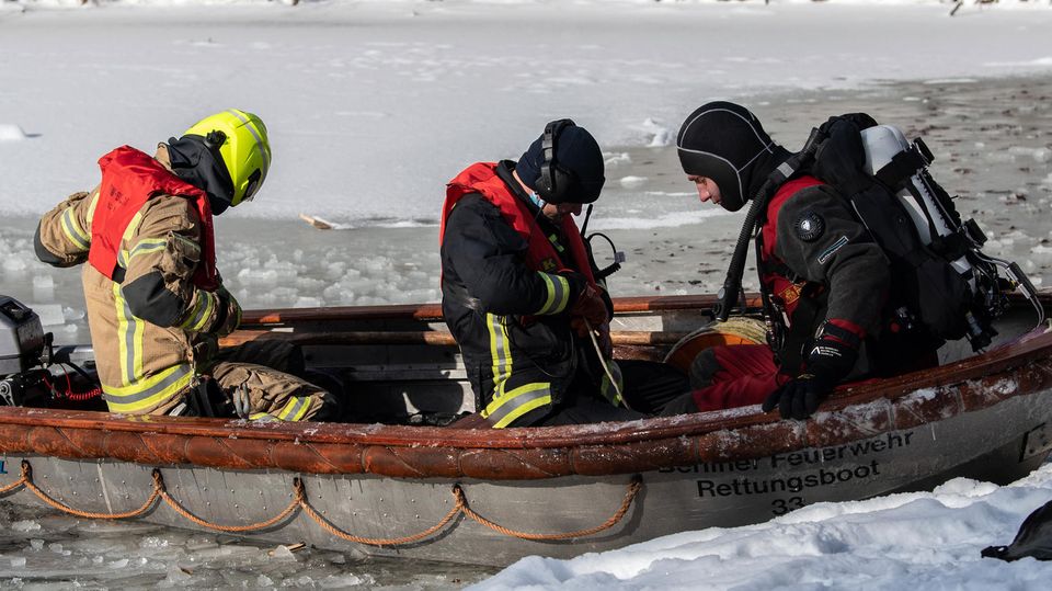 Zwei Personen in Feuerwehr-Ausrüstung und ein Taucher sitzen in einem Aluminium-Boot mit Außenborder auf einem zugefrorenen See