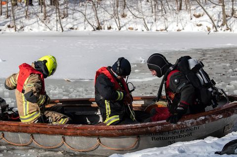 Zwei Personen in Feuerwehr-Ausrüstung und ein Taucher sitzen in einem Aluminium-Boot mit Außenborder auf einem zugefrorenen See