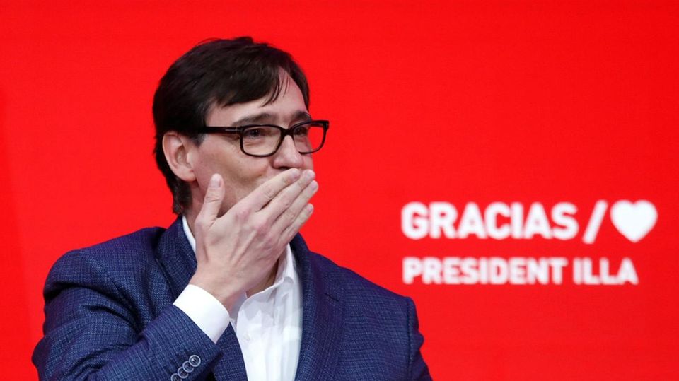 Salvador Illa Sozialisten Katalonien Wahl Ein Mann in blauem Anzug steht auf einem ganz in roten Podium und wirft Luftküsse ins Publikum. Er trägt eine Hornbrille
