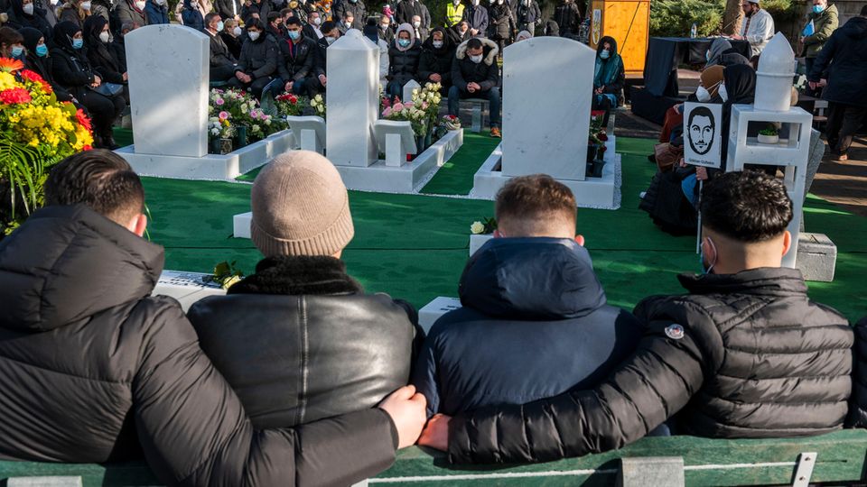 Hanau Gedenken Terror Opfer Friedhof Jahrestag Anschlag Von hinten sind vier junge Männer auf Stühlen zu sehen. Sie sitzen mit Blick auf vier Grabsteine und umarmen einander