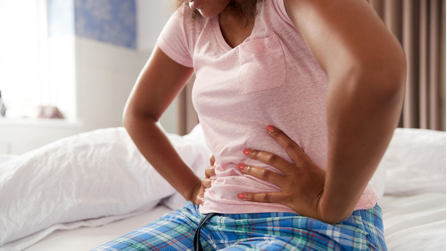Eine Frau im Schlafanzug sitzt auf dem Bett und drückt die Hände auf den Bauch