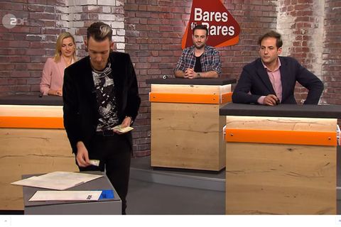Fabian Kahl steht im Bares für Rares Studio in Pulheim, neben ihm sitzen seine Kollegen
