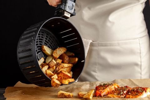 Heißluftfritteuse: Frau kippt fertige Kartoffelspalten aus einem Frittierkorb auf Backpapier