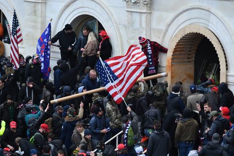 Eine Menschenmenge mit US- und Trump-Flaggen drängt bewaffnet gegen ein weißes Gebäude vor