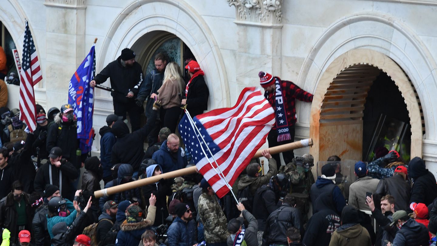 Eine Menschenmenge mit US- und Trump-Flaggen drängt bewaffnet gegen ein weißes Gebäude vor