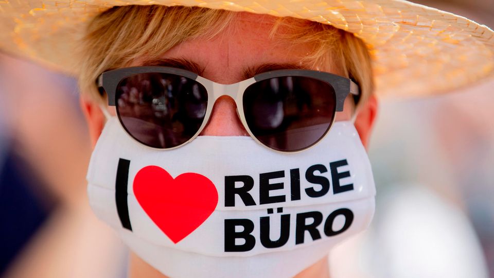 Eine Frau mit Sonnenbrille trägt eine Maske mit der Aufschrift "I love Reisebüro"
