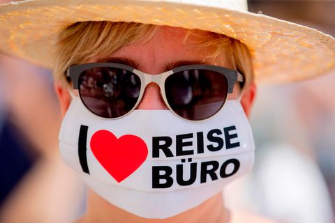 Eine Frau mit Sonnenbrille trägt eine Maske mit der Aufschrift "I love Reisebüro"