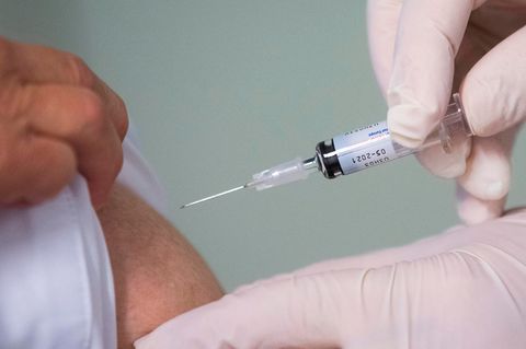 Eine rechte Hand in weißem Gummihandschuh ist kurz davor, eine Spritze in den Oberarm eines Mannes zu stechen