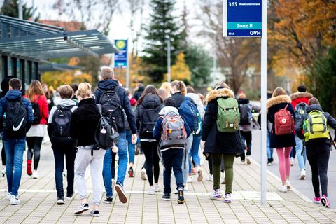 Schülerinnen und Schüler gehen mit ihren Schulrucksäcken von einer Bushaltestelle zu ihrer Schule