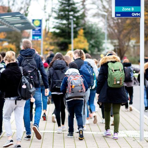 Schülerinnen und Schüler gehen mit ihren Schulrucksäcken von einer Bushaltestelle zu ihrer Schule