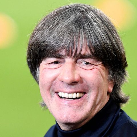In dunkelblauem Rollkragen-Pulli steht Jogi Löw am Rand eines Fußball-Platzes und lacht. Der Rasen im Hintergrund ist unscharf