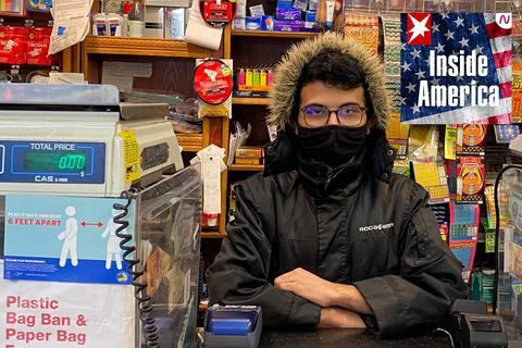 Ein dunkelhaariger junger Mann sitzt in schwarzer Winterjacke und mit verschränkten Armen hinter dem Tresen eines kleinen Ladens