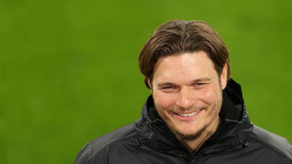 Ein Mann mit dunkelblondem Seitenscheitel grinst, während er auf einem Fußballplatz interviewt wird