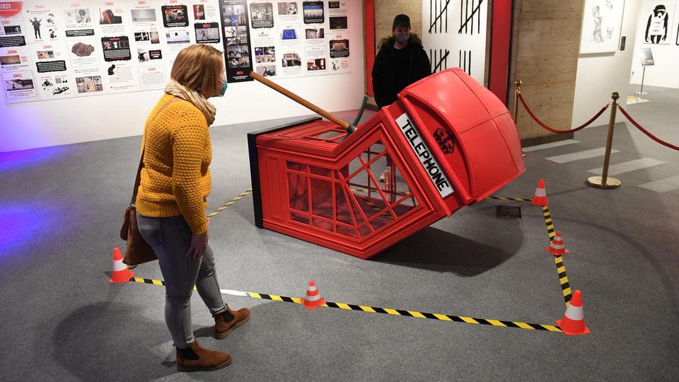 Leicht gebeugt betrachtet eine junge Frau in senfgelbem Wollpulli eine am Boden liegende, geknickte rote Telefonzelle von Banksy