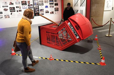 Leicht gebeugt betrachtet eine junge Frau in senfgelbem Wollpulli eine am Boden liegende, geknickte rote Telefonzelle von Banksy