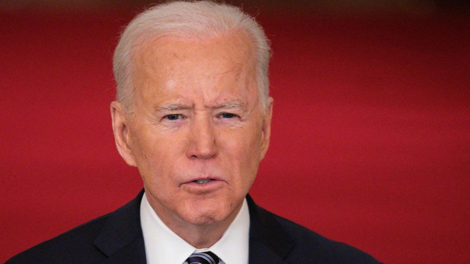 Joe Biden Vor einem roten Hintergrund steht ein älterer weißer Mann in Anzug und Krawatte. Er trägt eine US-Flagge am Revers