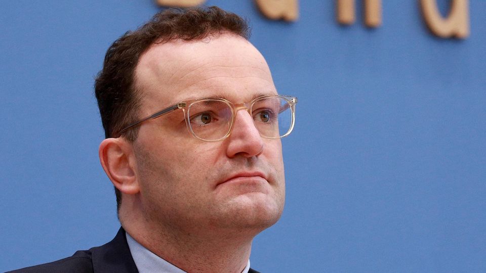 Ein Mann mit braunen Locken und durchsichtigem Brillengestell sitzt im Anzug vor einer blauen Wand