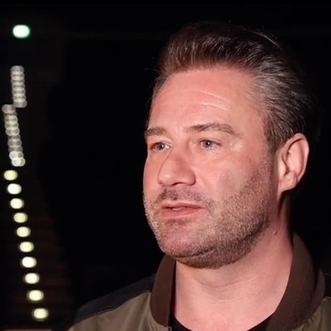 Ein Mann mit dunkelblonder Tolle steht in einer dunklen Arena, in der nur sein Gesicht ausgeleuchtet ist