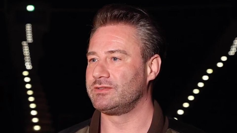 Sasha Ein Mann mit dunkelblonder Tolle steht in einer dunklen Arena, in der nur sein Gesicht ausgeleuchtet ist