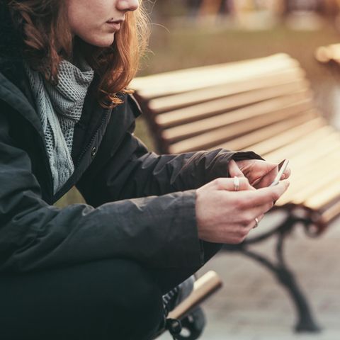 Junge Frau sitzt alleine auf einer Bank und schaut auf ein Smartphone.