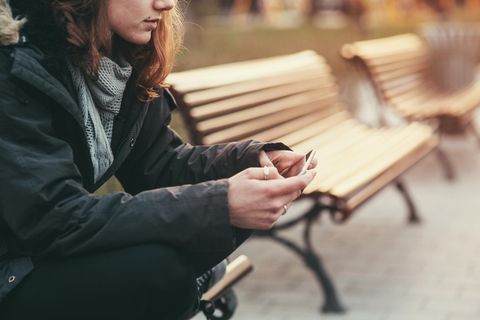 Junge Frau sitzt alleine auf einer Bank und schaut auf ein Smartphone.