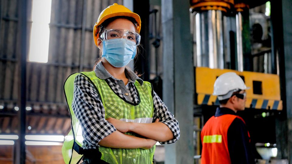 Arbeiterin mit hygienischer Maske steht selbstbewusst in einer Halle - mit ihrem Mitarbeiter als Hintergrund
