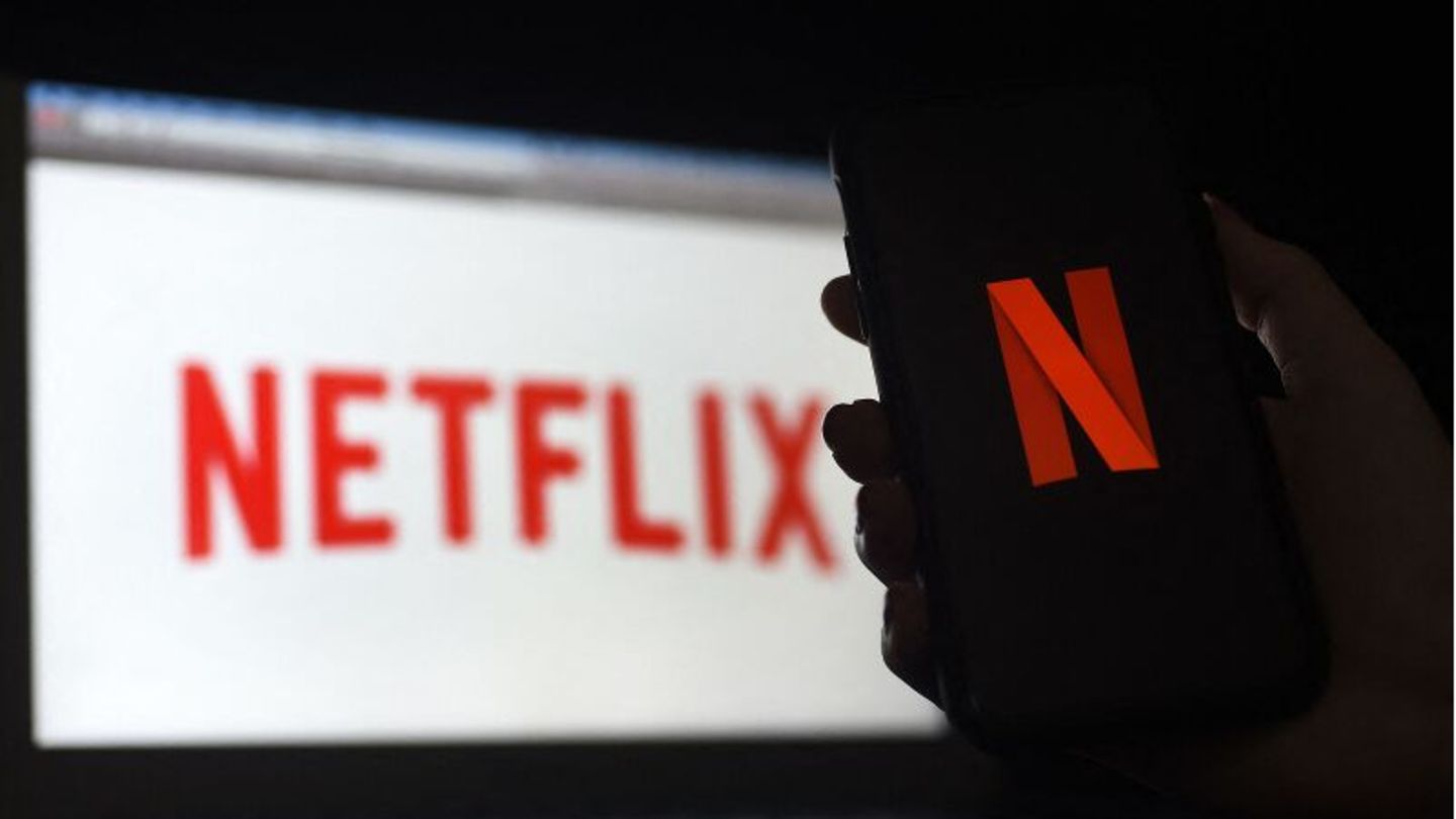 Eine Hand hält ein Smartphone mit dem Netflix-Bildschirm vor einem Laptop, auf dem ebenfalls ein Netflix-Logo zu sehen ist