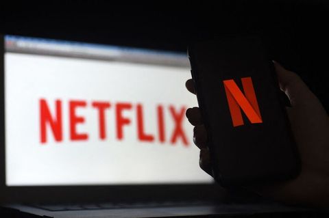 Eine Hand hält ein Smartphone mit dem Netflix-Bildschirm vor einem Laptop, auf dem ebenfalls ein Netflix-Logo zu sehen ist