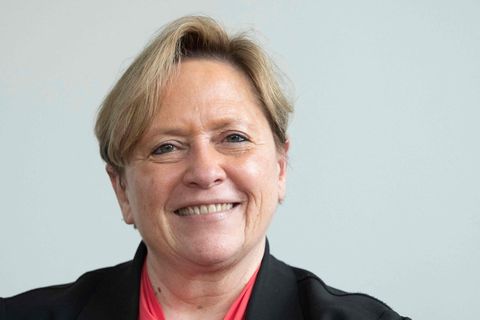 Eine ältere weiße Frau mit blondem Seitenscheitel, rotem Halstuch und schwarzem Blazer lächelt in die Kamera
