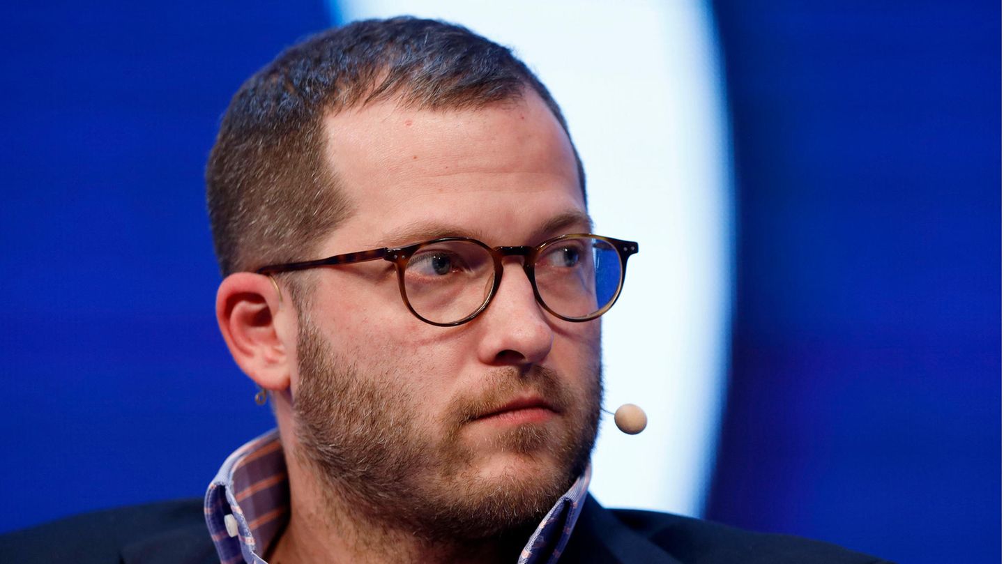 Ein Mann mit kurzen, braunen Haaren, Drei-Tage-Bart und Hornbrille sitzt mit zur Seite gedrehtem Kopf vor einer blau-weißen Wand