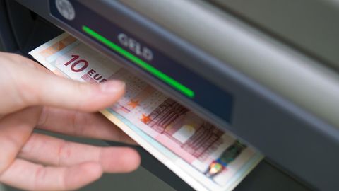 Eine Hand greift nach Geldscheinen, die aus einem Geldautomaten ausgeworfen wurden