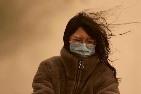 Eine junge Chinesin mit langen schwarzen Haaren fährt mit OP-Maske durch den Sandsturm, der den Himmel gelb einfärbt