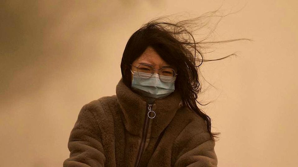 Eine junge Chinesin mit langen schwarzen Haaren fährt mit OP-Maske durch den Sandsturm, der den Himmel gelb einfärbt