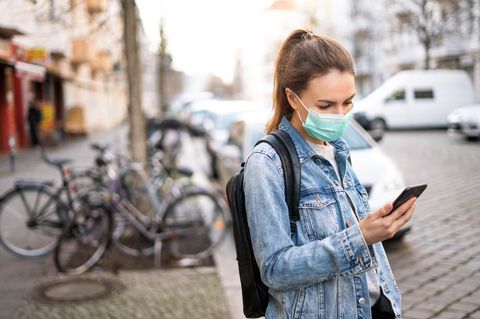 Frau mit Alltagsmaske steht an einer Straße und schaut auf ihr Handy