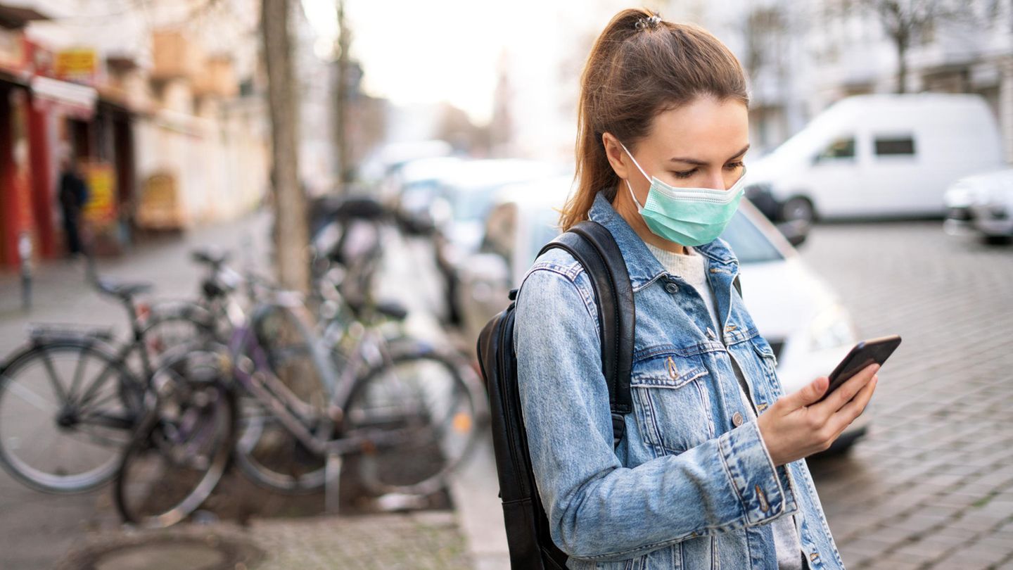 Frau mit Alltagsmaske steht an einer Straße und schaut auf ihr Handy