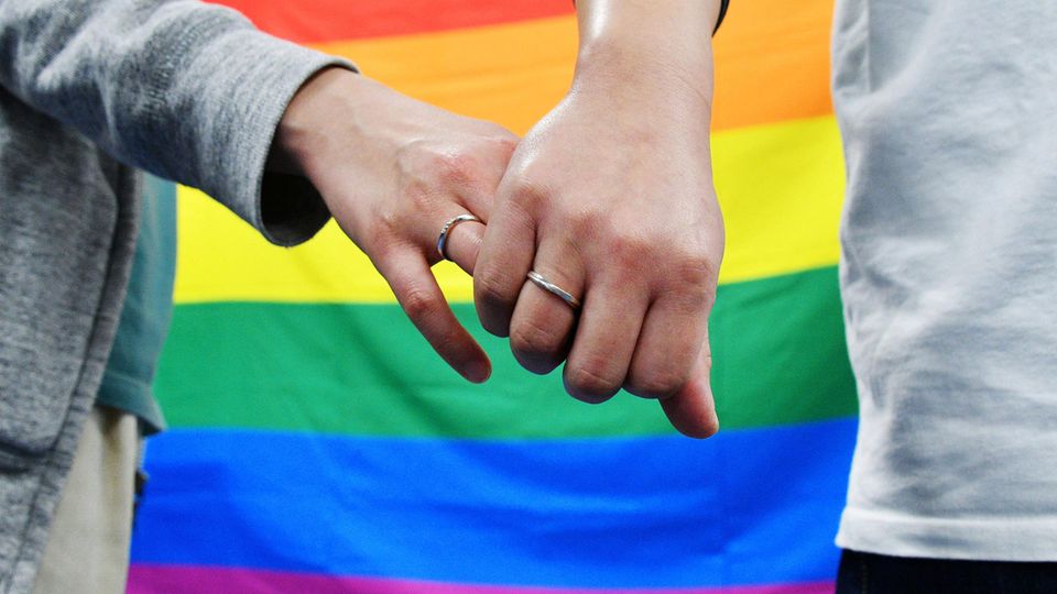 Ein gleichgeschlechtliches Paar hält sich vor einer Regenbogenflagge an den Händen