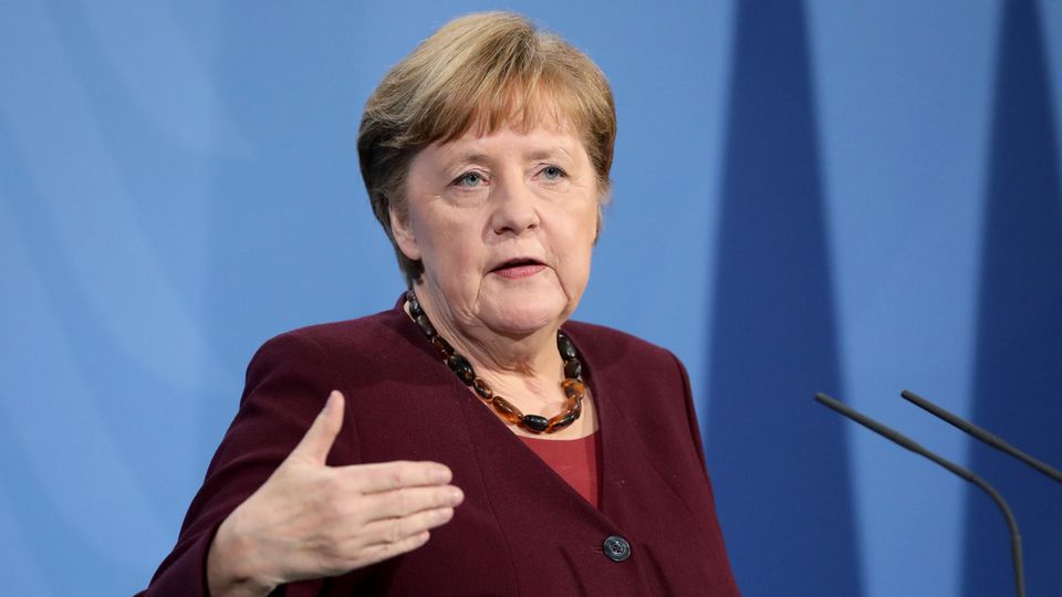 Angela Merkel steht in weinrotem Blazer vor einer blauen Wand, spricht in zwei Mikros und gestikuliert mit rechts