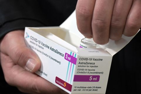 Eine rechte Männerhand hält einen weißen Karton mit Astrazeneca-Impfstoff, die linke zieht eine Ampulle heraus