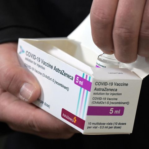 Eine rechte Männerhand hält einen weißen Karton mit Astrazeneca-Impfstoff, die linke zieht eine Ampulle heraus