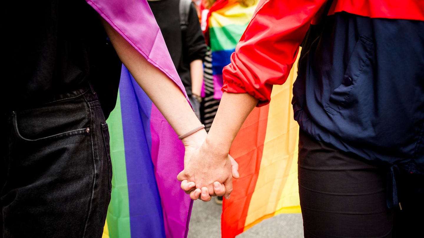 Ein homosexuelles Paar hält Hände vor einer Regenbogenflagge.