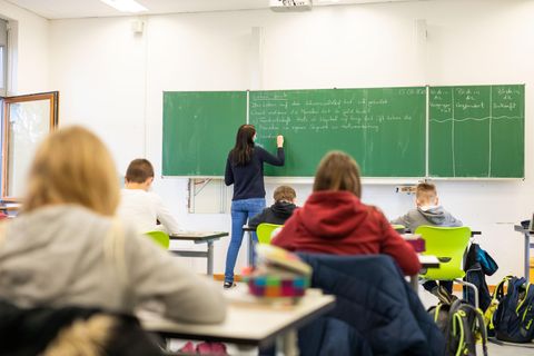 Schüler sitzen im Unterricht, während eine Lehrerin an die Tafel schreibt